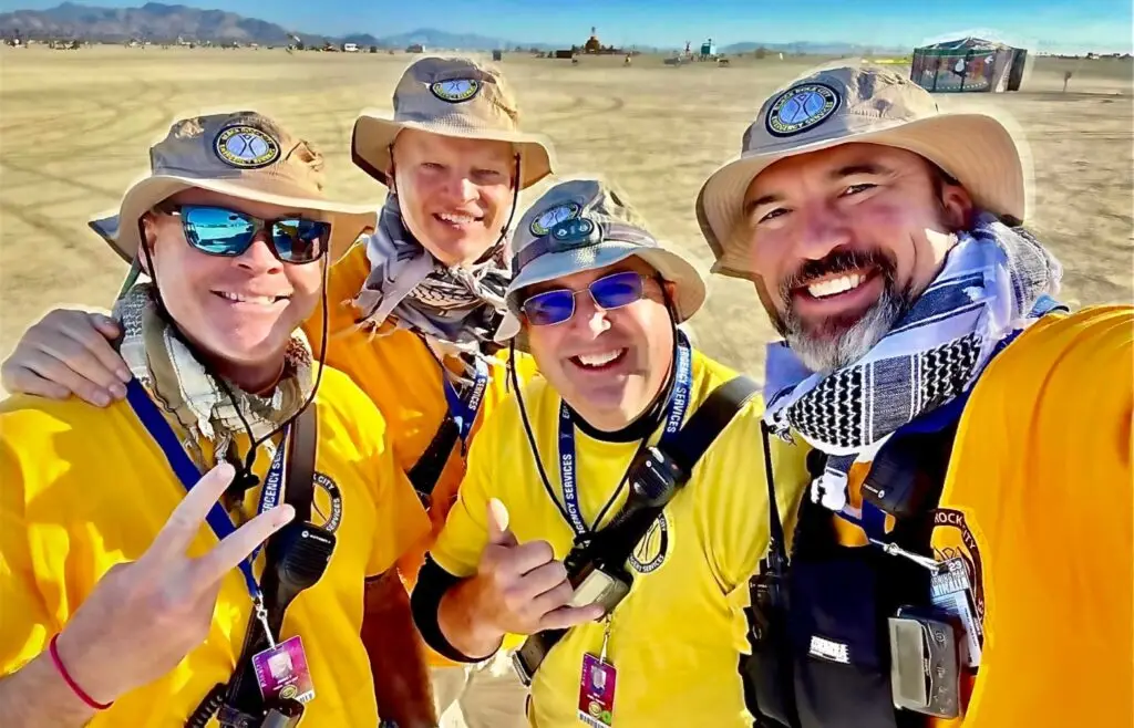Four friends smiling outdoors on a sunny day, wearing hats and yellow shirts.